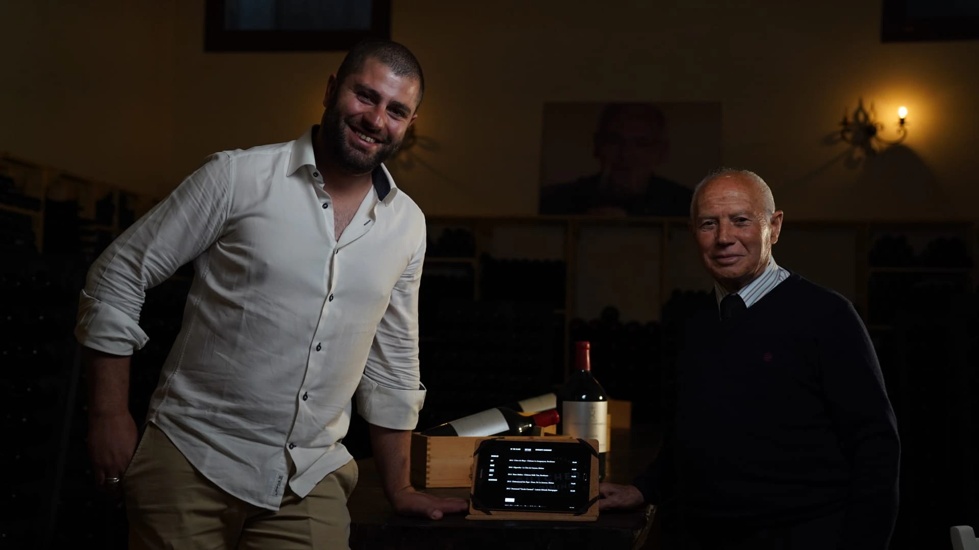 Twee mannen lachen in de wijnkelder van Baglio del Cristo di Campobello, met een tablet met de wijnkaart en een kist flessen tussen hen in.